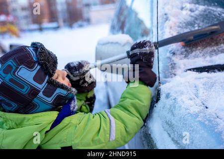 Simpatico ragazzo caucasico pulisce l'auto dalla neve con una spazzola e un raschietto, aiutando suo padre nell'inverno nevoso. Il ragazzino carino rimuove la neve Foto Stock