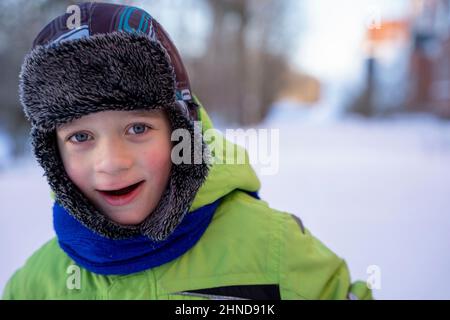 Divertente ragazzo caucasico di 5 anni vestito con caldi abiti invernali si erge sulla neve e guarda la macchina fotografica sorridente. Passeggiate invernali nel fresco Foto Stock