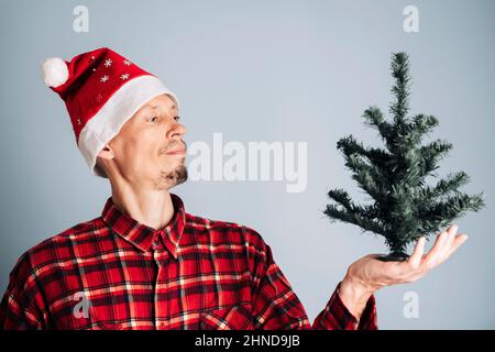 Una foto ravvicinata di un giovane uomo in una camicia rossa a quadri e un cappellino di San Nicola. Un uomo con capelli lunghi legati in un nodo tiene un albero di Natale sul suo braccio. Foto Stock