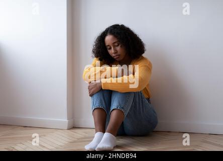 Tutta la lunghezza di giovane donna nera che soffre di depressione, seduta sul pavimento e abbracciando le ginocchia a casa, spazio copia Foto Stock