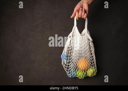 La mano di una donna tiene una borsa a corda con una bottiglia d'acqua, un arancio e mele verdi su uno sfondo scuro testurizzato, il concetto di prodotto senza rifiuti Foto Stock