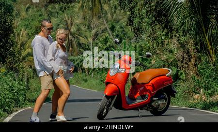 Due donne turistiche caucasiche uomo danza vicino scooter. Coppia d'amore su moto rossa in abiti bianchi, occhiali da sole sulla foresta strada percorso viaggio. Strada da ballo. Noleggio moto. Asia Thailandia giro turismo. Foto Stock
