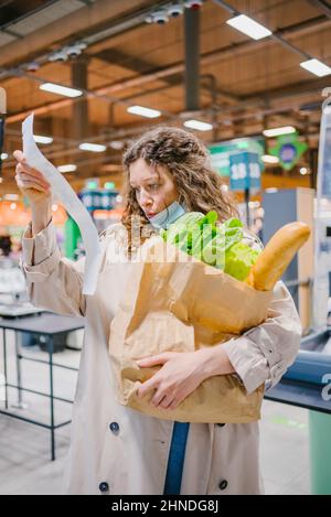 La giovane donna è sorpresa spiacevolmente mentre osserva in un controllo della carta in un supermercato della drogheria e tiene una borsa della spesa. Aumento dei prezzi dei prodotti alimentari. Foto Stock