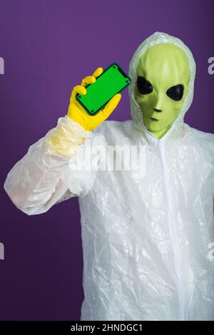 Alieno verde in tuta bianca mostra un telefono con uno schermo verde su sfondo viola. Foto Stock