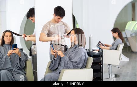 Ragazza cliente utilizzando il telefono durante il taglio dei capelli da professionale parrucchiere Foto Stock