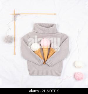 Tre gelati di filati e coni di cialde su un pullover grigio lavorato a maglia, aghi in legno da maglieria su sfondo bianco. Lavorazione a maglia, hobby e concep fatti a mano Foto Stock