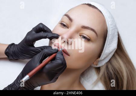 Giovane donna con trucco permanente sulle labbra in un salone di cosmetologia. Tatuaggio permanente per il trucco. Tracciare una linea con una matita rossa a labbro Foto Stock