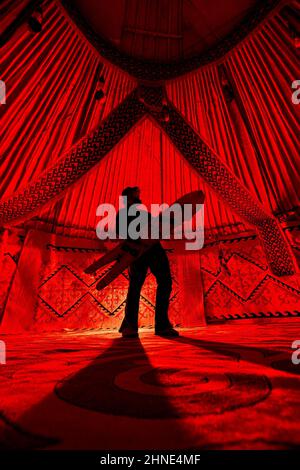 Silhouette di uomo che tiene split snowboard come la chitarra all'interno di Yurt nomade casa contro tappeto con motivi etnici illuminati da luce rossa interno. Backco Foto Stock