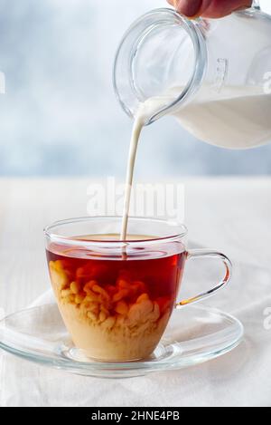 Versare il latte in una tazza di tè nero su un tavolo di legno bianco Foto Stock