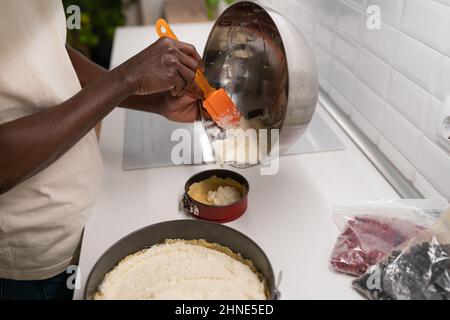 Uomo africano che fa cheesecake con bacche surgelate in cucina. Primo piano del processo di cottura fatto in casa Foto Stock