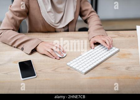 visualizzazione ritagliata della donna utilizzando la tastiera e il mouse del computer mentre si lavora vicino allo smartphone con schermo vuoto Foto Stock