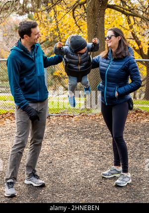 Giovani genitori che spingono il ragazzo di due anni su un'altalena da parco giochi della città; Philadelphia; Pennsylvania; USA Foto Stock