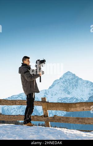 Uomo videografo che realizza riprese utilizzando una fotocamera montata su un dispositivo stabilizzatore gimbal. Neve e montagne sullo sfondo. Foto Stock