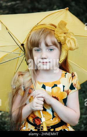 bella ragazza dai capelli rossi con arco giallo e ombrello giallo. Una ragazza di 11 anni guarda la fotocamera e sorride. Foto Stock