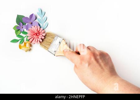 Womans mano che tiene la spazzola davanti ai fiori di carta su sfondo bianco. Arte della carta e concetto artigianale. Stagione primaverile Foto Stock