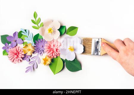 Womans mano che tiene la spazzola davanti ai fiori di carta su sfondo bianco. Arte della carta e concetto artigianale. Stagione primaverile e design floreale Foto Stock