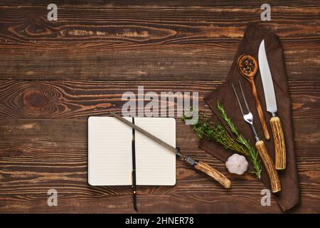 Forchetta, coltello e affilatrice vintage su tavola in legno accanto al notebook. Apparecchi per barbecue e open notepad, blogging di cibo e concetto di menu. Foto Stock