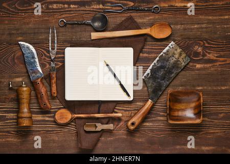 Vista dall'alto di forchetta, coltello e berlina vintage su un tavolo di legno accanto al notebook. Apparecchi per barbecue e blocco note aperto, blogging di cibo e menu conce Foto Stock