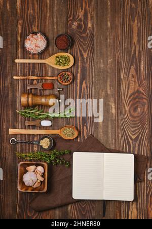 Vista dall'alto del blocco note aperto per ricette accanto a cucchiai da cucina e ciotole con varie spezie, erbe e condimenti. Concetto di cucina di piccante e piccante Foto Stock