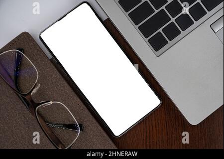 Vista dall'alto del telefono sul tavolo vicino a computer portatile, blocco note e occhiali con schermo bianco. Foto Stock