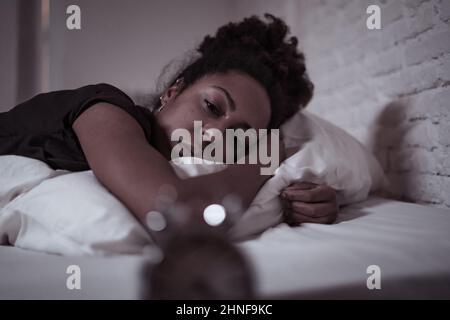 Insonnia e disperata bella afro americana donna svegliarsi di notte non in grado di dormire guardando l'orologio che soffre di insonnia in disturbi del sonno e. Foto Stock