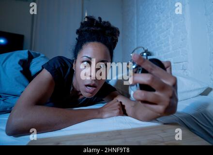 Insonnia e disperata bella afro americana donna svegliarsi di notte non in grado di dormire guardando l'orologio che soffre di insonnia in disturbi del sonno e. Foto Stock