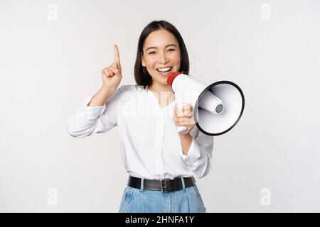 Sorridendo ragazza asiatica felice che parla in megaphone e che punta in su, annunciando promo di sconto, mostrando la pubblicità in cima, in piedi su sfondo bianco Foto Stock