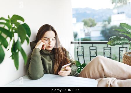 Giovane bella donna su un balcone utilizzando il telefono cellulare chatting.Unhappy giovane donna controllare il suo smartphone al mattino. Triste annoiato persona adulta seduta nel balcone usando il suo telefono come distrazione. Foto Stock