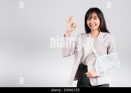 Braccio rotto. Business donna fiducioso sorriso rotto braccio dopo incidente indossare stecca braccio per il trattamento mostra OK segno, felice Asian femmina imbracatura sostegno mano Foto Stock