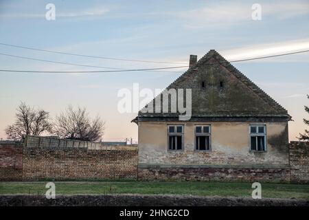 Foto di una fattoria abbandonata in Vojvodina, in Serbia, con la facciata della sua casa principale gravemente danneggiata e decadente. Foto Stock