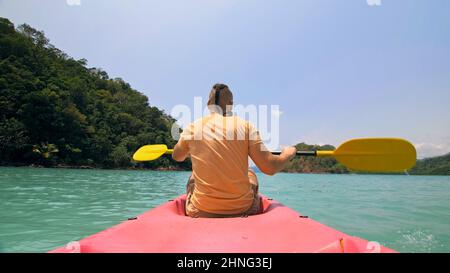 Giovane uomo con occhiali da sole e cappello ranghi rosa canoe di plastica lungo il mare contro verdi isole collinari con giungle selvatiche. Viaggiare in paesi tropicali. Il ragazzo forte sta navigando in kayak nell'oceano, vista posteriore. Foto Stock