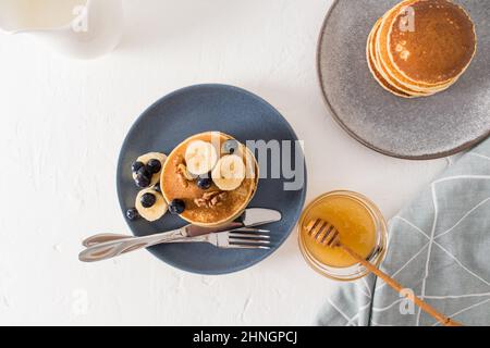 una pila di pancake su un piatto scuro con noci e fette di banana. miele in una ciotola con un cucchiaio per migliorare il gusto. vista dall'alto. sfondo bianco Foto Stock