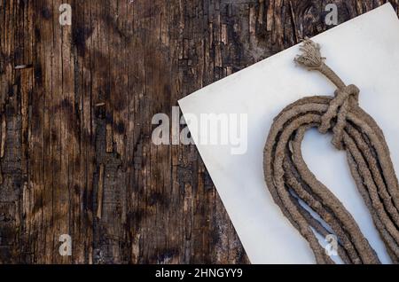 Un rotolo di vecchia corda e un pezzo di carta bianco su sfondo di legno scuro. Fune sporca e usurata. Superficie di legno incrinata con crepe e trucioli. Selettivo f Foto Stock