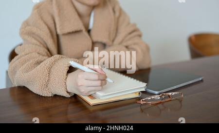 Una donna millennial che scrive giù il suo programma di studio nel suo diario, elencando le sue idee sul blocco note a spirale, scrivendo le note sul taccuino, seduto nel bar Foto Stock