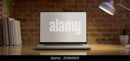 Un computer portatile portatile bianco schermo mockup sotto la luce dalla lampada da tavolo su tavolo in legno su pareti di mattoni rossi nella moderna sala d'ufficio vintage. 3d rende Foto Stock