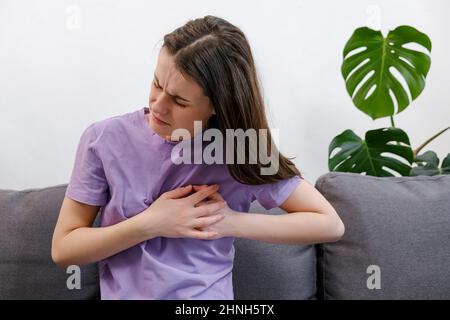 Turbata giovane caucasica donna sensazione dolore toccare petto avendo attacco di cuore, triste infelice ragazza preoccupata soffre di mal di cuore sedersi sul divano Foto Stock