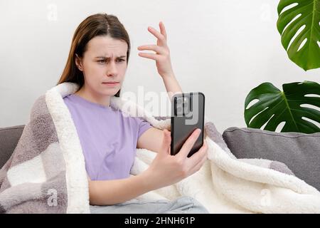 Cattive notizie sullo schermo. Confuso frustrata giovane donna coperta plaid sedersi sul divano guardando smartphone vedere importante chiamata persa. Disperato millennial la Foto Stock