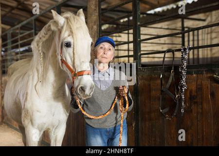 Lavoratrice stalla femminile anziata che conduce il cavallo bianco dalla briglia in fienile Foto Stock