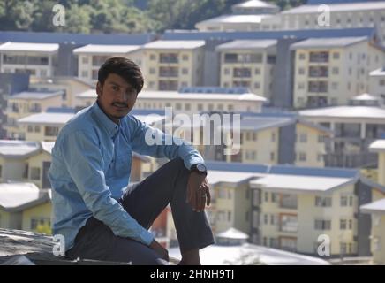 Un ragazzo giovane che guarda la macchina fotografica seduta fuori con edifici in background con spazio copia Foto Stock