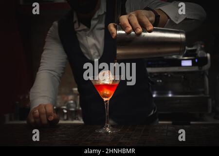 un barista professionista prepara un drink nel bicchiere di martini con un liquido molto appariscente di colore arancio, un barista maschile elegantemente vestito Foto Stock