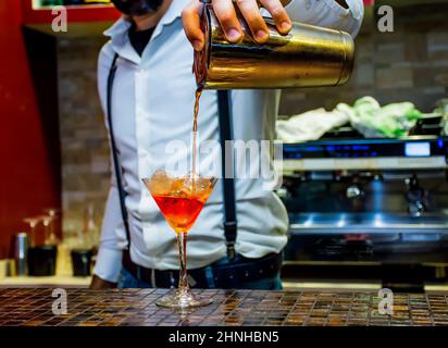 un barista professionista prepara un drink nel bicchiere di martini con un liquido molto appariscente di colore arancio, un barista maschile elegantemente vestito Foto Stock