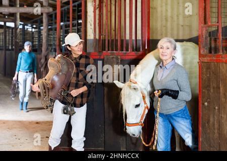 Donna asiatica che tiene la sella mentre anziana femmina stabile custode che porta cavallo fuori dalla stalla Foto Stock