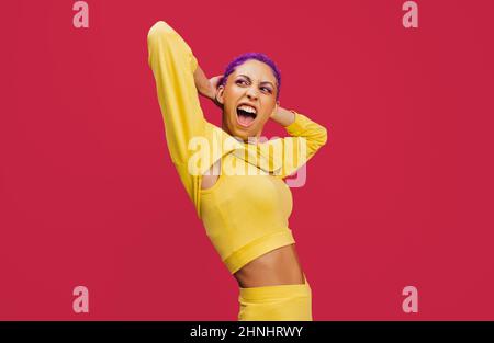 Giovane donna elegante in piedi con le mani dietro la testa in uno studio. Giovane donna alla moda che indossa trucco con capelli viola. Giovane donna bein Foto Stock