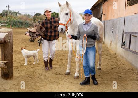 Anziana stalla donna che conduce cavallo a cavallo all'arena di equitazione all'aperto, mentre donna asiatica che tiene sella Foto Stock