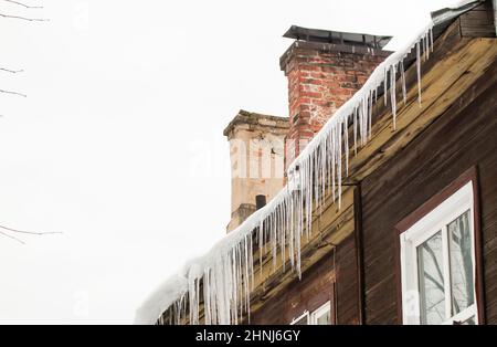 Le ghiere lisce e trasparenti pendono dal bordo del tetto. Sullo sfondo del muro di legno della vecchia casa. Grandi cascate, anche belle file. Nuvoloso giorno d'inverno, luce tenue. Foto Stock