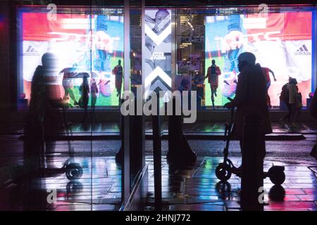 Londra, UK, 16 febbraio 2022: Su Oxford Street, un enorme schermo a LED si riflette dai marciapiedi e lungo una strada laterale in una serata piovosa. Gli acquirenti e i pendolari con ombrelloni o e-scooters sono in perfetto contrasto con le luci brillanti. L'economia si sta ribellando dopo la pandemia e la fiducia dei consumatori sembra tornare nonostante le preoccupazioni per il costo della vita di crisi. Anna Watson/Alamy Live News Foto Stock