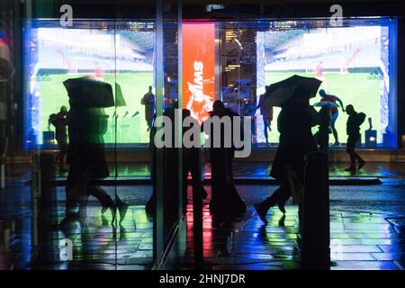 Londra, UK, 16 febbraio 2022: Su Oxford Street, un enorme schermo a LED si riflette dai marciapiedi e lungo una strada laterale in una serata piovosa. Gli acquirenti e i pendolari con ombrelloni o e-scooters sono in perfetto contrasto con le luci brillanti. L'economia si sta ribellando dopo la pandemia e la fiducia dei consumatori sembra tornare nonostante le preoccupazioni per il costo della vita di crisi. Anna Watson/Alamy Live News Foto Stock
