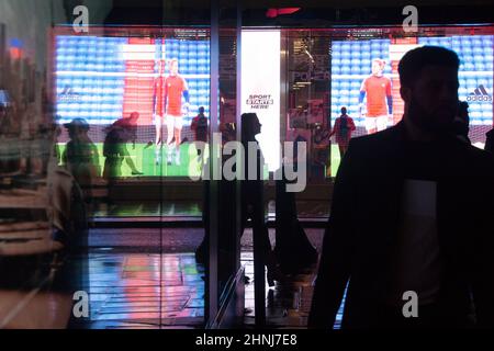 Londra, UK, 16 febbraio 2022: Su Oxford Street, un enorme schermo a LED si riflette dai marciapiedi e lungo una strada laterale in una serata piovosa. Gli acquirenti e i pendolari con ombrelloni o e-scooters sono in perfetto contrasto con le luci brillanti. L'economia si sta ribellando dopo la pandemia e la fiducia dei consumatori sembra tornare nonostante le preoccupazioni per il costo della vita di crisi. Anna Watson/Alamy Live News Foto Stock