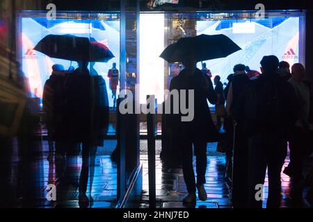 Londra, UK, 16 febbraio 2022: Su Oxford Street, un enorme schermo a LED si riflette dai marciapiedi e lungo una strada laterale in una serata piovosa. Gli acquirenti e i pendolari con ombrelloni o e-scooters sono in perfetto contrasto con le luci brillanti. L'economia si sta ribellando dopo la pandemia e la fiducia dei consumatori sembra tornare nonostante le preoccupazioni per il costo della vita di crisi. Anna Watson/Alamy Live News Foto Stock