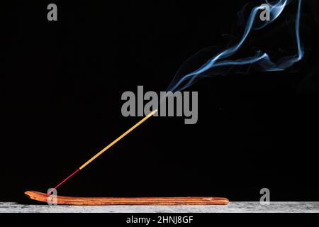 Bastone ardente di incenso si erge su un basamento di legno isolato su uno sfondo nero. Foto Stock